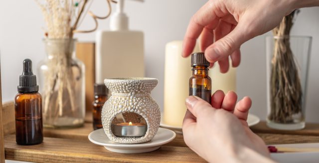 Hand is holding a bottle of essential oil and opening the lid. There is an aroma lamp, candles and cosmetics on the table. Concept of aromatherapy, relaxing atmosphere, stress management and rituals